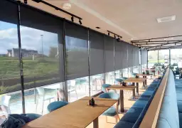 Nowoczesne wnętrze restauracji z widokiem na wodę, osłonięte eleganckimi roletami termicznymi na wymiar.
