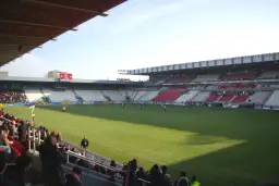 Widok na murawę i trybuny stadionu Cracovii. Na boisku trwa mecz, a trybuny zapełniają się kibicami.