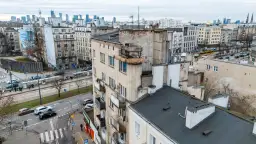 Widok na zaniedbany budynek mieszkalny z balkonami i panoramę miasta w tle.