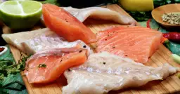 Stek z łososia kontra polędwica z dorsza. Surowe kawałki ryb, przyprawione pieprzem, leżą obok limonki, cytryny i ziół.