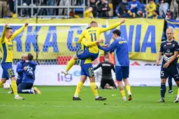 Piłkarze w żółto-niebieskich strojach świętują zwycięstwo na boisku, jeden z nich jest niesiony przez kolegę.
