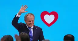 Donald Tusk macha do tłumu na tle niebieskiego ekranu z czerwono-białym sercem, symbolem Koalicji Obywatelskiej.
