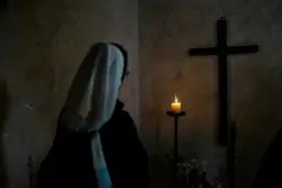 Zakonnica w białym welonie patrzy na płonącą świecę i krzyż w ciemnym pomieszczeniu.