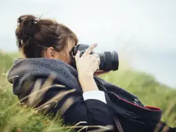Kobieta z włosami spiętymi w kok leży na trawie, robiąc zdjęcie profesjonalną lustrzanką do vlogów.