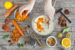 Ręce dodają startą marchewkę do ciasta. Czy gotowana marchewka jest zdrowa? Składniki na ciasto marchewkowe: marchew, pomarańcza, orzechy.