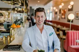 Uśmiechnięty mężczyzna w jasnoniebieskiej marynarce siedzi przy barze. Zastanawiasz się, ile zarabia manager hotelu?