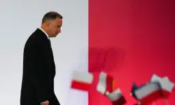 Andrzej Duda w czarnym garniturze, pochylony, idzie na tle czerwonej ściany z flagami. Czy to prawdziwe powody dla których Andrzej Duda zapowiedział serię wet do ustaw budżetowych?