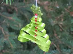 Urocza choinka z zielonej wstążki, ozdobiona czerwonymi koralikami, wisi na gałęziach iglaka.