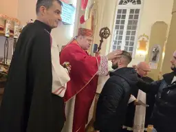 Biskup w czerwonej szacie udziela sakramentu bierzmowania młodemu mężczyźnie w kościele.