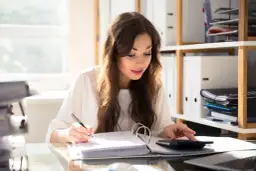 Kobieta w biurze pisze w segregatorze i używa kalkulatora.