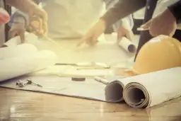 Zespół analizuje plany, dyskutując, ile trwa podział działki. Na stole leżą zrolowane projekty, miarka i żółty kask.
