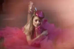 Urocza sesja glamour: blondynka w różowej tiulowej sukni i błyszczącej beretce, z makijażem oczu w tym samym kolorze, leży na ziemi.