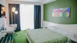A modern hotel room with a green bedspread, green carpet, and a colorful abstract art piece on a green wall.
