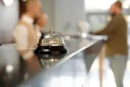 Dzwonek hotelowy na ladzie, symbol usług uzupełniających. W tle niewyraźne postacie personelu.
