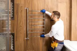 Mężczyzna w niebieskich rękawiczkach sprawdza grzejnik łazienkowy. Dowiedz się, jak zakręcić grzejnik łazienkowy.