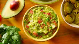 Miseczka guacamole z pomidorami i cebulą, obok awokado, kolendra i plasterki jalapeno.