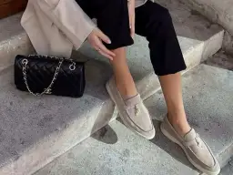 Kobieta w beżowych mokasynach i czarnych spodniach siedzi na schodach obok czarnej torebki Chanel.