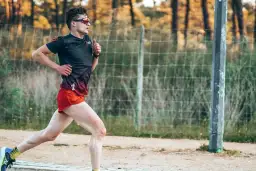 Mężczyzna w sportowym stroju biegnie, ćwicząc jak zwiekszyc kadencje w bieganiu. W tle siatka i drzewa.