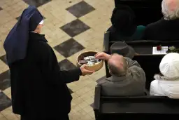 Zakonnica w czarnym habicie trzyma koszyk z pieniędzmi, do którego starszy mężczyzna wkłada banknot.