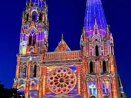 Katedra w Chartres nocą, rozświetlona kolorowymi projekcjami. Architektura gotycka w odcieniach niebieskiego i pomarańczowego.