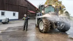 Mężczyzna myje traktor myjką gorącowodną przed stodołą.