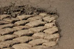 Zbliżenie na dziurę w asfalcie z widocznymi kamieniami i krawędzią asfaltu.