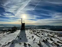 Szczyt Babiej Góry zimą. Krzyż i kamienna konstrukcja na ośnieżonym szczycie. W tle rozległa panorama gór i niebo z chmurami. Szlaki Babiej Góry w śniegu.