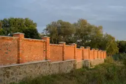 Solidny płot murowany z cegły i kamienia, z ozdobnymi słupkami, otacza teren porośnięty zielenią.