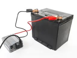 Ładowanie akumulatora 100Ah: prostownik podłączony do czarnego akumulatora za pomocą czerwonych i czarnych klem.