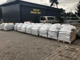 Palety z workami materiału budowlanego przed budynkiem sklepu drogowego.