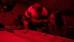 W ciemni fotograficznej, przy czerwonym świetle, mężczyzna i dziewczyna pracują przy powiększalniku i kuwetach.