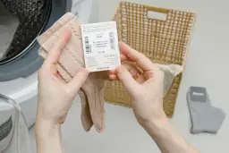 Dłonie trzymające beżowe skarpetki z metką obok koszyka i pralki.