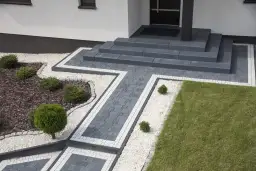 Nowoczesny podjazd z szarej kostki brukowej, schody i zieleń.