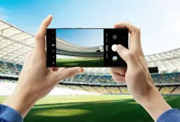 Dwie dłonie trzymają smartfon Samsung S23, robiąc zdjęcie pustego stadionu piłkarskiego.