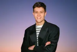 Młody mężczyzna w czarnej marynarce i pasiastym t-shircie z łańcuszkiem na szyi, na tle nieba.