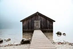 Drewniana szopa nad jeziorem, z pomostem prowadzącym do drzwi. Kompozycja otwarta przykłady.