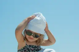 Dziecko w okularach przeciwsłonecznych i białej czapce UV dla dzieci, z rękami na głowie, na tle błękitnego nieba.