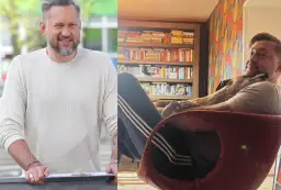 Marcin Prokop w swetrze i z psem na kolanach na tle regału z książkami.