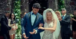 Para młoda wychodzi z kościoła, obsypana płatkami. Panna młoda w białej sukni, pan młody w granatowym garniturze.