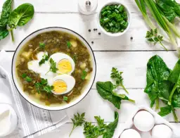 Zupa szczawiowa z jajkiem i śmietaną, otoczona świeżym szpinakiem, szczypiorkiem i jajkami.