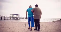 Mężczyzna z laską, wspierany przez drugiego, idzie po plaży. Może to symbolizować wsparcie w walce o odszkodowanie za stwardnienie rozsiane.