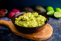 Miseczka guacamole z kawałkami cebuli i pomidorów, na drewnianej desce, w tle awokado i limonki.