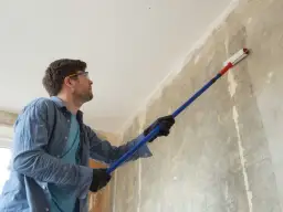 Mężczyzna w okularach ochronnych i rękawiczkach nakłada grunt na ścianę wałkiem. To pierwszy krok, jak zrobić grunt do ścian, by przygotować je do malowania.