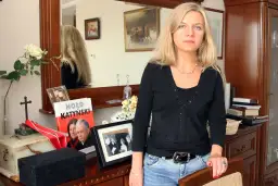 Kobieta z blond włosami w czarnej bluzce i jeansach, obok książka "Hołd Katyński" i ramka ze zdjęciem.