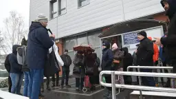 Grupa ludzi stoi przed budynkiem spółdzielni mieszkaniowej w deszczu, niektórzy pod parasolem.