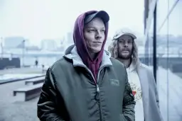 Dwóch mężczyzn w kapturach i czapkach, wyglądają jakby szli na jakieś seanse. Inni ludzie w tle.