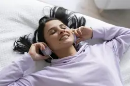 Kobieta w słuchawkach leży na łóżku, zrelaksowana. Słucha muzyki na dobry sen, uśmiechając się lekko.