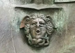 Brązowa rzeźba anioła z opaską na oczach i ustach, z wężami u dołu.
