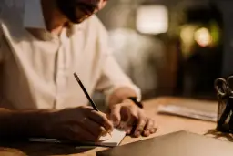 Mężczyzna w białej koszuli pisze ołówkiem w notatniku. Tworzy wartościowy branded content, pracując nad ważnym projektem.