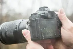 Dłoń trzyma aparat z obiektywem, pokryty kroplami deszczu. To była trudna, ale udana sesja w deszczu.